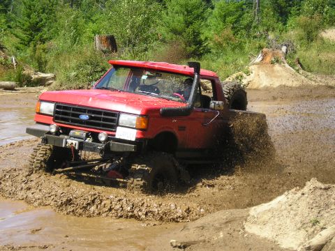 1989 Ford Bronco - A red Ford Bronco navigates a muddy off-road course, kicking up a spray of mud.