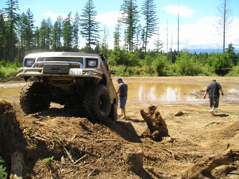 1989 Ford Bronco - A heavily modified truck navigates a muddy obstacle course with two onlookers nearby.