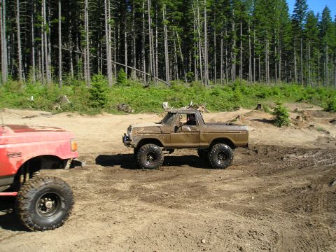 1989 Ford Bronco - Two off-road trucks navigate a muddy path through a wooded area.