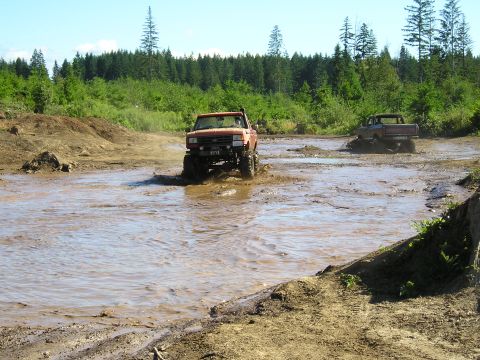 1989 Ford Bronco - Two pickup trucks navigate a muddy off-road course with trees in the background.