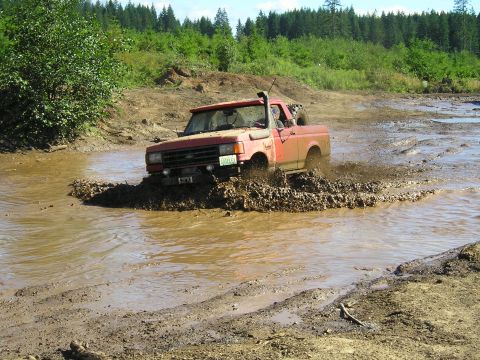1989 Ford Bronco - A red 1987 Ford Bronco navigates a challenging muddy trail, kicking up water and dirt.