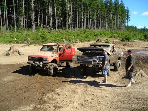 1989 Ford Bronco - Two modified trucks are parked in a dirt lot with trees behind them.