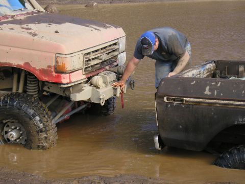 1989 Ford Bronco - A man inspects two off-road vehicles submerged in muddy water, one appearing to be a 1989 Ford Bronco.