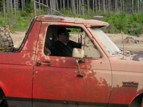 1989 Ford Bronco - A driver sits in a heavily muddied red Ford Bronco, its body style indicating a 1987 model year.