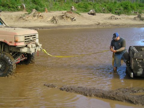 1989 Ford Bronco - Two trucks stuck in a muddy water hole, one being pulled out with a yellow strap.