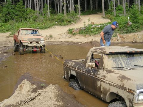 1989 Ford Bronco - Two off-road trucks are stuck in the mud and water, with one vehicle attempting to tow the other.