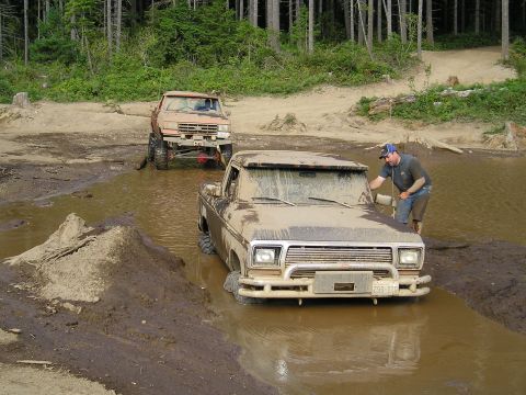 1989 Ford Bronco - A man cleans the windshield of a mud-splattered pickup truck stuck in a muddy puddle, with another truck behind it.