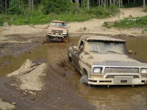 1989 Ford Bronco - Two off-road vehicles, a pickup truck and an SUV, navigate a muddy water obstacle on a wooded trail.