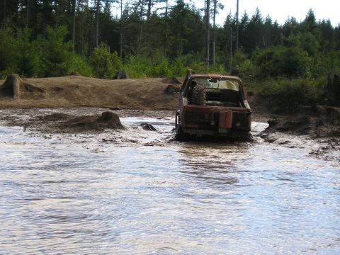 1989 Ford Bronco - A red off-road vehicle navigates through a muddy, water-filled terrain with dense trees lining the horizon.