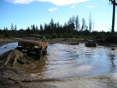 1989 Ford Bronco - Two trucks navigate a muddy water crossing in a wooded area.