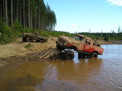 1989 Ford Bronco - Two off-road trucks navigate a muddy trail with trees in the background under a clear blue sky.