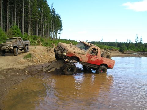 1989 Ford Bronco - Two modified pickup trucks tackle a challenging muddy water crossing in a wooded area.