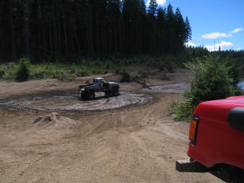 1989 Ford Bronco - Two vehicles on a muddy off-road course with trees and a blue sky.