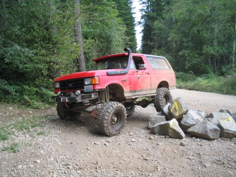 1989 Ford Bronco - A red Ford Bronco navigates an obstacle course on a dirt road surrounded by trees.
