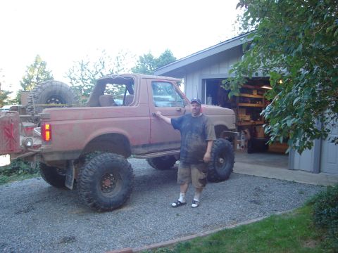 1989 Ford Bronco - A man stands beside a heavily modified, older model Ford Bronco, parked in front of a suburban garage.