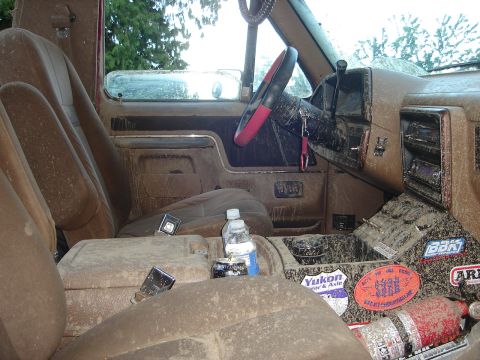 1989 Ford Bronco - The interior of a 1989 Ford Bronco is covered in mud, with stickers adorning the center console and a red steering wheel cover visible.