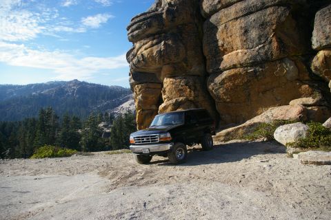 1996 Ford Bronco - A black 1996 Ford Bronco is parked on a dirt road, with a massive rock formation towering over it and a scenic mountain range in the distan…