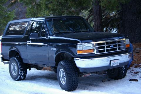 1996 Ford Bronco - A black 1996 Ford Bronco is parked in a snowy, wooded area, its lifted suspension and off-road tires.