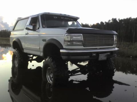 1995 Ford Bronco - A lifted white Ford Bronco sits in shallow water with a forest in the background.