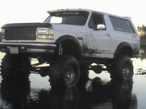 1995 Ford Bronco - This lifted Ford Bronco, modified for off-roading, is shown in shallow water with mud splatters on its body.
