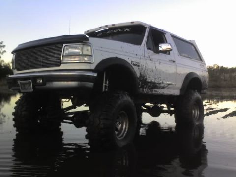 1995 Ford Bronco - A lifted white Ford Bronco with large tires sits in shallow water, covered in mud.