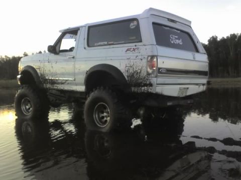 1995 Ford Bronco - This lifted 1995 Ford Bronco is captured mid-action, navigating through a muddy terrain with its tires splashing.