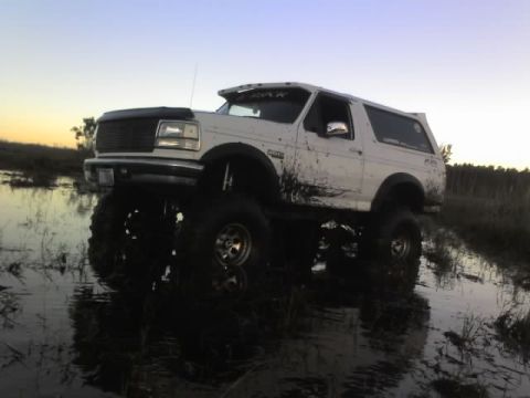 1995 Ford Bronco - A white Bronco navigates through a muddy, watery terrain under a clear sky.