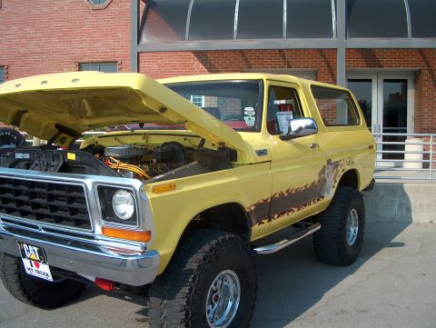 1978 Ford Bronco - A yellow 1978 Ford Bronco is parked outside a brick building with its hood open, revealing the engine.