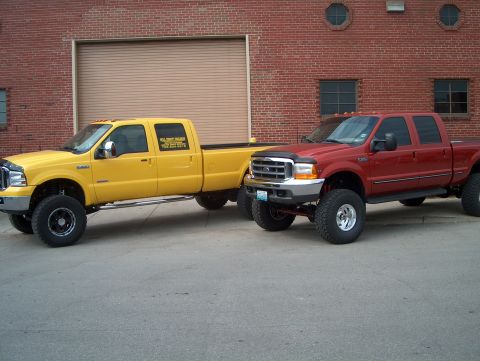 1978 Ford Bronco - Two heavy-duty pickup trucks, one yellow and one red, are parked side-by-side in front of a brick building.