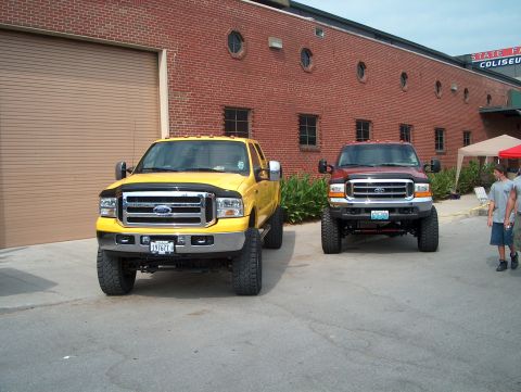 1978 Ford Bronco - Two lifted Ford trucks, one yellow and one red, are parked outside a brick building.