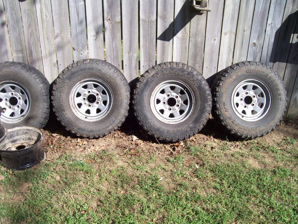 Four Mastercraft Courser M/T tires on silver steel wheels leaning against a wood fence