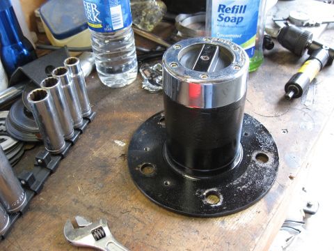 1994 Ford Bronco - A freshly painted black front wheel hub with a chrome-bodied locking hub assembly installed, sitting upright on a cluttered wooden workbenc…