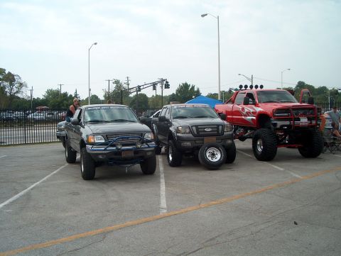 1978 Ford Bronco - A trio of heavily modified pickup trucks, including a red one with flame decals and a gray one with spare tires, are parked together in an…