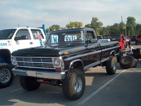 1978 Ford Bronco - A lifted black pickup truck is parked in a lot with other vehicles and people.