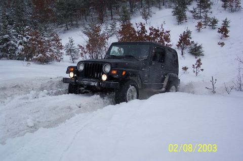 1978 Ford Bronco - A black Jeep Wrangler navigates a snowy, wooded terrain, kicking up snow as it drives.