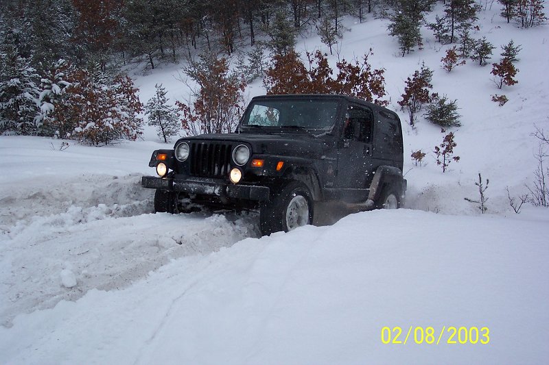 black Jeep Wrangler driving through deep snow on a wooded hill