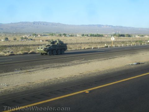 1990 Ford Bronco - green eight-wheeled military armored vehicle moves along a desert highway with mountains in the distance.