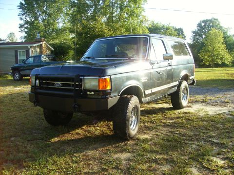 1990 Ford Bronco - This Ford Bronco is parked in a grassy yard next to a house, with a pickup truck visible in the background.