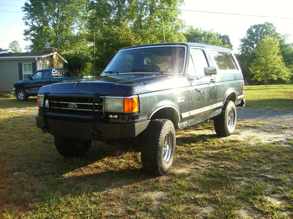 Dark green Ford Bronco with aftermarket bumper and 4x4 badging. A green pickup truck is parked in the background