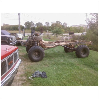 1977 Ford F-150 - A truck chassis with large tires and engine is parked on a grassy hill, with another truck partially visible in the foreground.