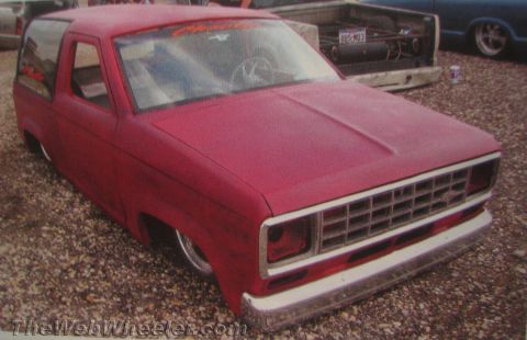 1990 Ford Bronco - lowered red two-door compact pickup in flat primer sits slammed on the gravel with a dropped suspension and chrome bumper.