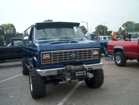 1978 Ford Bronco - Blue lifted Ford Econoline van on a 4x4 chassis, photographed from the 3/4 front in a paved show lot. It has a chrome front bumper with a m…