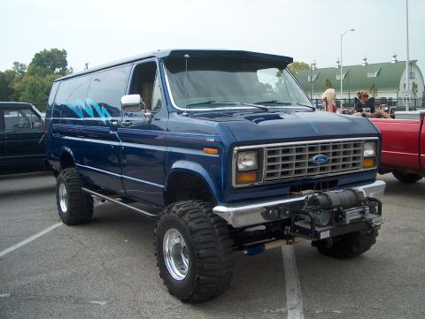 1978 Ford Bronco - Three-quarter front view of a lifted 4x4 blue Ford Econoline van in a parking lot, running mud-terrain tires on polished slotted wheels wit…