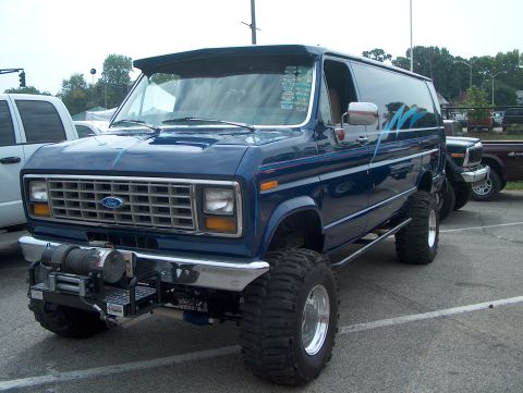 1978 Ford Bronco - A dark blue lifted Ford Econoline full-size van shown from the front three-quarter angle, riding on large mud-terrain tires with chrome whe…