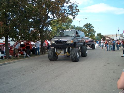 1978 Ford Bronco - Dark gray Ford Econoline van built as a monster truck, rolling down a closed street in a parade with spectators on the sidewalks. It rides…
