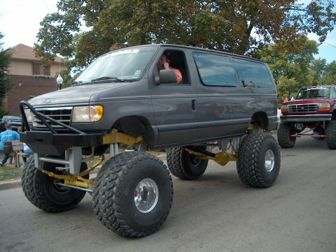 1978 Ford Bronco - Three-quarter front view of a grey Ford Econoline van built as a monster truck, riding on huge terra-style flotation tires with polished wh…