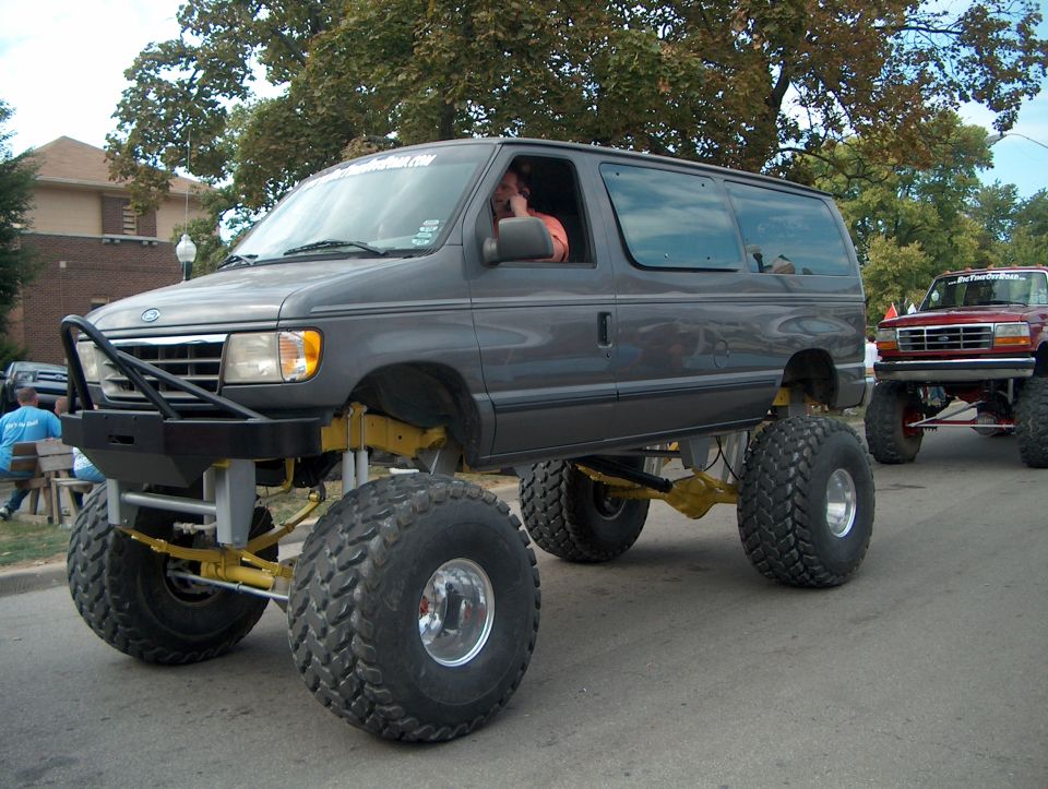 Grey lifted Ford Econoline van on huge terra tires with yellow axles at a street show