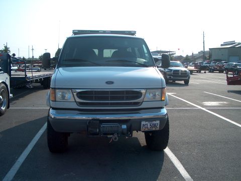 1978 Ford Bronco - Head-on front view of a silver Ford Econoline van lifted on 4x4 running gear, parked in a paved lot. A winch with hook and yellow strap is…