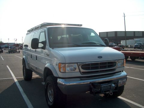 1978 Ford Bronco - White Ford Econoline full-size van converted to four-wheel drive, parked in an asphalt lot and shot from the front three-quarter angle. It…