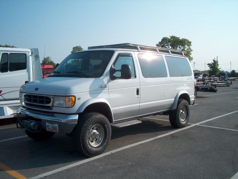 1978 Ford Bronco - Silver Ford E-450 Super Duty van converted to 4x4, viewed from the 3/4 front in a parking lot. It wears a roof rack, running boards, BFGood…